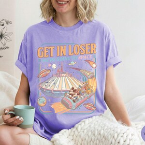 Pode incluir: Camiseta violeta com um gr&aacute;fico retr&ocirc; que diz "Get in Loser, We're Going to Space". O gr&aacute;fico apresenta um passeio de parque tem&aacute;tico espacial com personagens de desenhos animados e discos voadores. A camiseta &eacute; feita de um material macio e confort&aacute;vel.