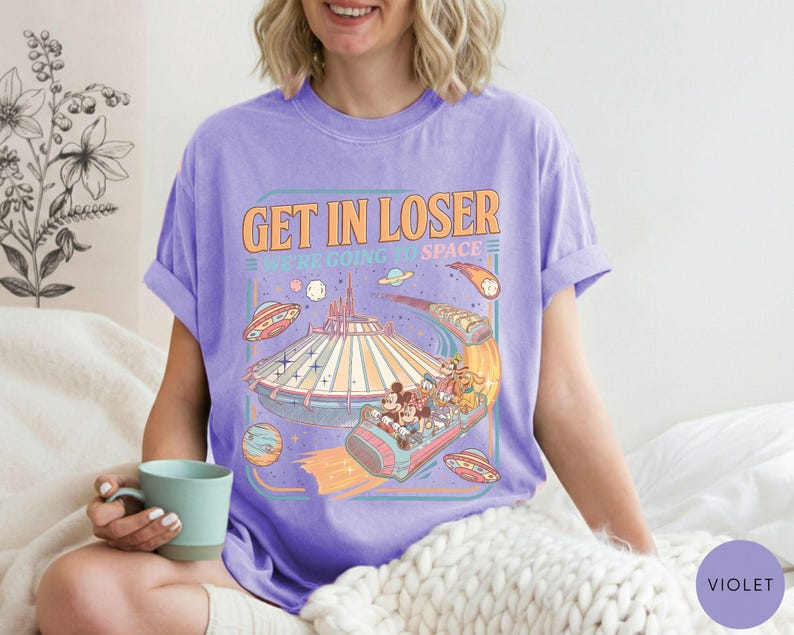 Pode incluir: Camiseta violeta com um gr&aacute;fico retr&ocirc; que diz "Get in Loser, We're Going to Space". O gr&aacute;fico apresenta um passeio de parque tem&aacute;tico espacial com personagens de desenhos animados e discos voadores. A camiseta &eacute; feita de um material macio e confort&aacute;vel.