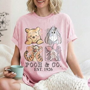 May include: Pink t-shirt featuring Winnie the Pooh, Eeyore, Tigger, and Piglet in a grid design. The shirt has the text "POOH & CO. EST. 1926" in black. The shirt is a light pink color.