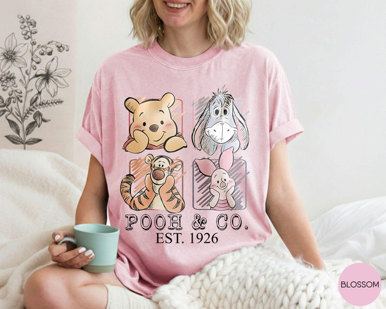 May include: Pink t-shirt featuring Winnie the Pooh, Eeyore, Tigger, and Piglet in a grid design. The shirt has the text "POOH & CO. EST. 1926" in black. The shirt is a light pink color.