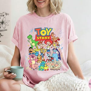 May include: A light pink t-shirt featuring a colourful graphic print of various Toy Story characters including Woody, Buzz Lightyear, Jessie, and Rex. The design includes the Toy Story logo in bold red and yellow letters, surrounded by playful scribbles.