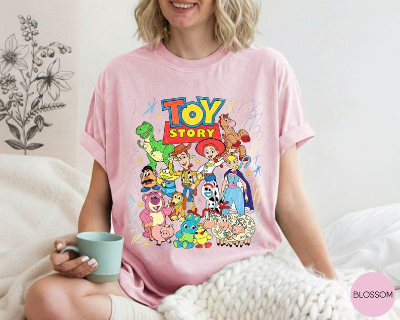 May include: A light pink t-shirt featuring a colourful graphic print of various Toy Story characters including Woody, Buzz Lightyear, Jessie, and Rex. The design includes the Toy Story logo in bold red and yellow letters, surrounded by playful scribbles.