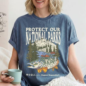 May include: Blue jean t-shirt with the text "PROTECT OUR NATIONAL PARKS" above a graphic of a mountain scene and a fish. The graphic includes trees, water, and a fish with orange and yellow fins. The text "Explore Responsibly" is below the graphic.