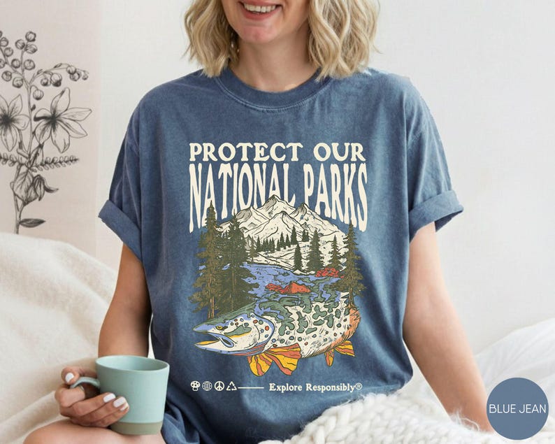 May include: Blue jean t-shirt with the text "PROTECT OUR NATIONAL PARKS" above a graphic of a mountain scene and a fish. The graphic includes trees, water, and a fish with orange and yellow fins. The text "Explore Responsibly" is below the graphic.