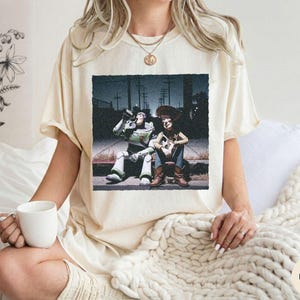 May include: An ivory-colored short-sleeve t-shirt featuring a graphic print of two characters, a space ranger and a cowboy, sitting on a curb at night. The shirt has a crew neckline and a relaxed fit, showcasing a detailed scene with dark tones.