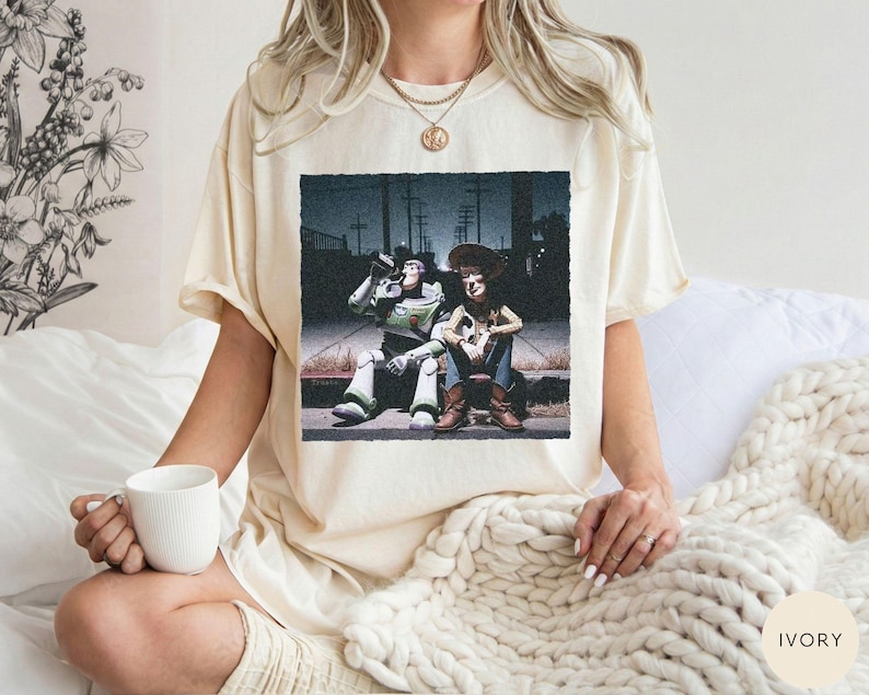 May include: An ivory-colored short-sleeve t-shirt featuring a graphic print of two characters, a space ranger and a cowboy, sitting on a curb at night. The shirt has a crew neckline and a relaxed fit, showcasing a detailed scene with dark tones.