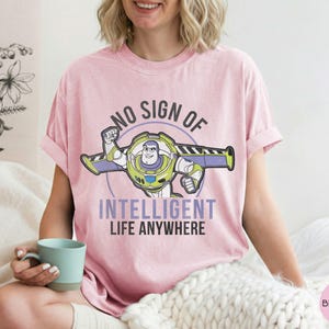 Puede incluir: Camiseta rosa claro con un dise&ntilde;o de Buzz Lightyear y el texto "NO SIGN OF INTELLIGENT LIFE ANYWHERE". El dise&ntilde;o incluye verde, morado y blanco. La camiseta es de estilo casual, ideal para el uso diario.