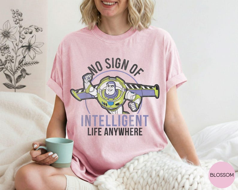 Puede incluir: Camiseta rosa claro con un dise&ntilde;o de Buzz Lightyear y el texto "NO SIGN OF INTELLIGENT LIFE ANYWHERE". El dise&ntilde;o incluye verde, morado y blanco. La camiseta es de estilo casual, ideal para el uso diario.