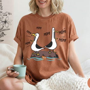 May include: A rust-coloured t-shirt with a cartoon illustration of two seagulls on a rock, with the word "MOM!" coming from them. A small cartoon fish is in the water below. The shirt is worn by a person.