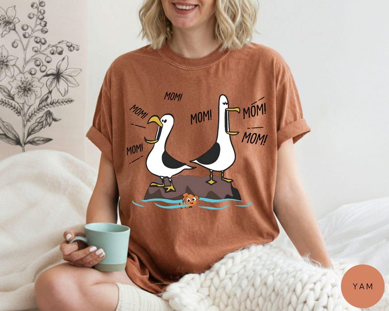 May include: A rust-coloured t-shirt with a cartoon illustration of two seagulls on a rock, with the word "MOM!" coming from them. A small cartoon fish is in the water below. The shirt is worn by a person.