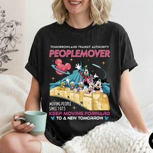 May include: Black t-shirt featuring a colorful graphic of the Tomorrowland Transit Authority PeopleMover ride with Mickey Mouse, Minnie Mouse, Goofy, Donald Duck, and Daisy Duck. The shirt includes the text "PEOPLEMOVER" and "KEEP MOVING FORWARD TO A NEW TOMORROW."