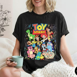 May include: A black t-shirt featuring a colourful graphic print of various Toy Story characters including Woody, Buzz Lightyear, Jessie, and Rex. The design includes the Toy Story logo in red and yellow, surrounded by white scribbled line art accents.