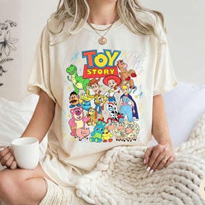 May include: An ivory-coloured t-shirt featuring a colourful graphic print of Toy Story characters including Woody, Buzz Lightyear, Jessie, and Rex. The design includes the text Toy Story in bold letters and various hand-drawn style scribbles.