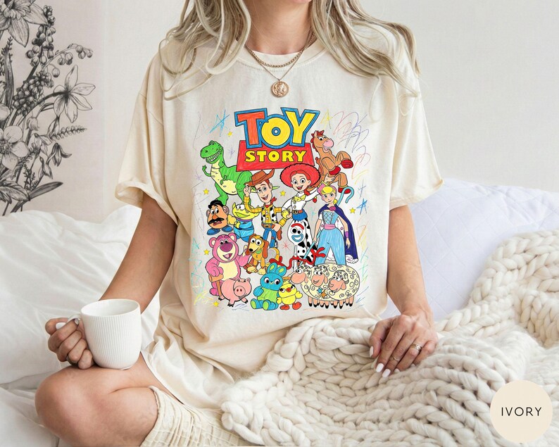 May include: An ivory-coloured t-shirt featuring a colourful graphic print of Toy Story characters including Woody, Buzz Lightyear, Jessie, and Rex. The design includes the text Toy Story in bold letters and various hand-drawn style scribbles.