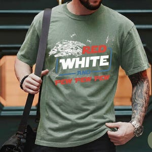 May include: An olive green short-sleeve t-shirt featuring a graphic of a spaceship and TIE fighters. The text reads RED WHITE AND PEW PEW PEW in a mix of red, white, and blue colors. The shirt has a crew neckline and a standard fit design.