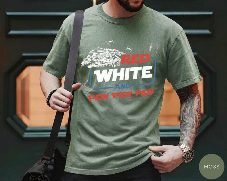 May include: An olive green short-sleeve t-shirt featuring a graphic of a spaceship and TIE fighters. The text reads RED WHITE AND PEW PEW PEW in a mix of red, white, and blue colors. The shirt has a crew neckline and a standard fit design.