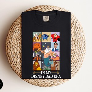 May include: Black t-shirt featuring a colorful grid of Disney character illustrations. The grid includes characters like Mr. Incredible, Dory, and King Triton. The text "IN MY DISNEY DAD ERA" is printed at the bottom. The shirt is displayed on a woven surface.