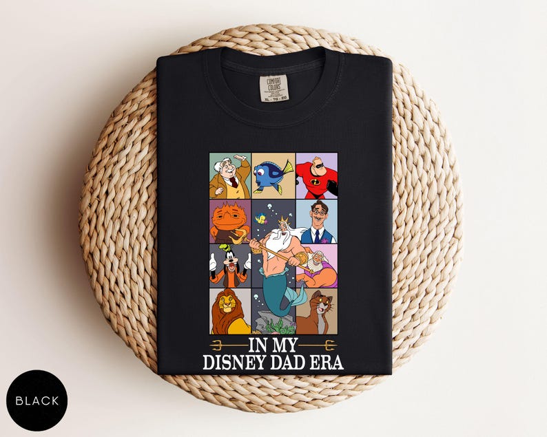 May include: Black t-shirt featuring a colorful grid of Disney character illustrations. The grid includes characters like Mr. Incredible, Dory, and King Triton. The text "IN MY DISNEY DAD ERA" is printed at the bottom. The shirt is displayed on a woven surface.