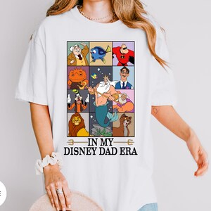 May include: White t-shirt featuring a colorful graphic of Disney characters in a grid pattern. The design includes characters like Mr. Incredible, Dory, and King Triton. The text "IN MY DISNEY DAD ERA" is printed below the grid.