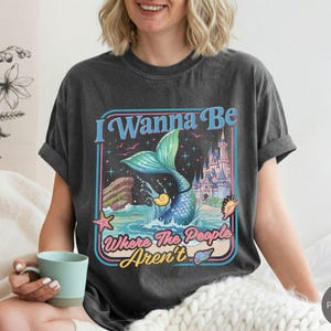 May include: Dark gray t-shirt with a mermaid graphic. The graphic features a mermaid with a blue tail, a castle, and the text "I Wanna Be Where The People Aren't" in a retro design.