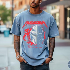 Op de afbeelding: Blauw spijkerbroekkleurig T-shirt met een rode en witte afbeelding van een Mandalorian helm en de tekst "MANDALORIAN" en "THIS IS THE WAY". Het shirt heeft korte mouwen en een relaxte pasvorm.