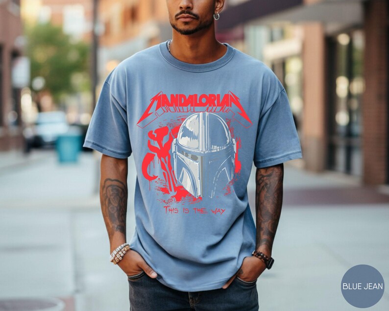 Op de afbeelding: Blauw spijkerbroekkleurig T-shirt met een rode en witte afbeelding van een Mandalorian helm en de tekst "MANDALORIAN" en "THIS IS THE WAY". Het shirt heeft korte mouwen en een relaxte pasvorm.