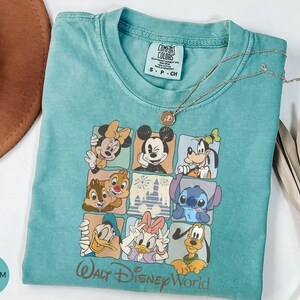 Może przedstawiać: Miętowa koszulka z nadrukiem przedstawiającym postacie Disneya, takie jak Myszka Miki i Myszka Minnie, Goofy, Kaczor Donald i Kaczka Daisy, Chip i Dale oraz Stitch. Napis "Walt Disney World" jest wydrukowany w kolorze złotym.