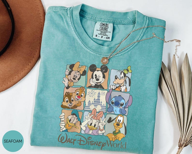 Może przedstawiać: Miętowa koszulka z nadrukiem przedstawiającym postacie Disneya, takie jak Myszka Miki i Myszka Minnie, Goofy, Kaczor Donald i Kaczka Daisy, Chip i Dale oraz Stitch. Napis "Walt Disney World" jest wydrukowany w kolorze złotym.