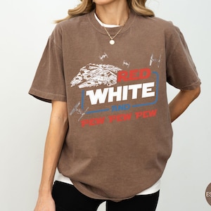 May include: A brown short-sleeve t-shirt featuring a white graphic of a spaceship and smaller fighter ships. The text reads RED WHITE AND PEW PEW PEW in red, white, and blue colors. The shirt has a crew neckline and a relaxed, loose-fitting cut.