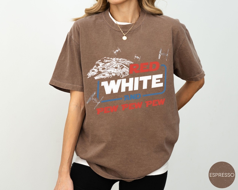 May include: A brown short-sleeve t-shirt featuring a white graphic of a spaceship and smaller fighter ships. The text reads RED WHITE AND PEW PEW PEW in red, white, and blue colors. The shirt has a crew neckline and a relaxed, loose-fitting cut.