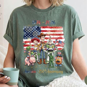 Pu&ograve; includere: T-shirt verde con una grafica colorata della bandiera americana, fuochi d'artificio e personaggi dei cartoni animati. La grafica include il testo "1776-2016" e "America 250th Anniversary".
