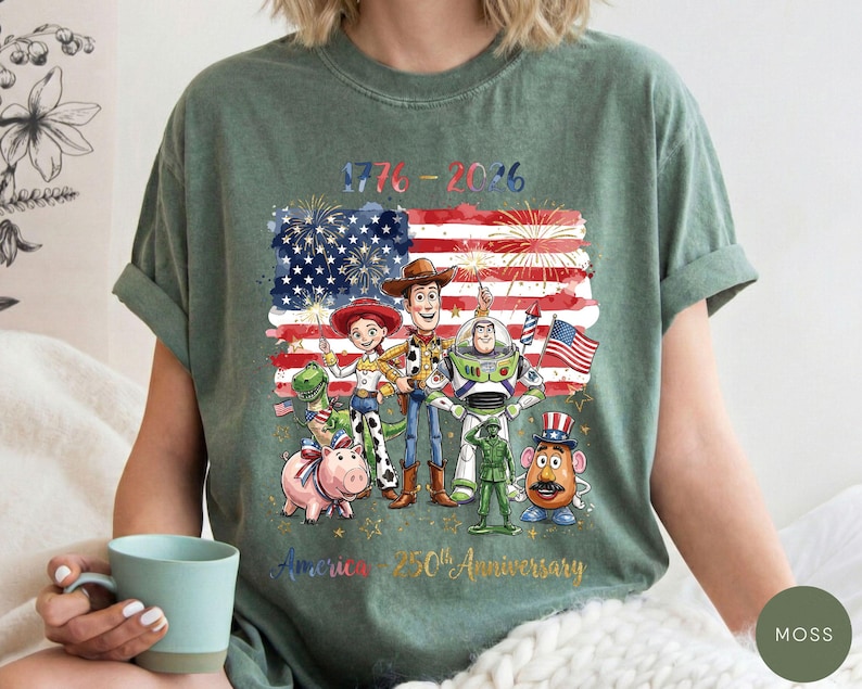 Pu&ograve; includere: T-shirt verde con una grafica colorata della bandiera americana, fuochi d'artificio e personaggi dei cartoni animati. La grafica include il testo "1776-2016" e "America 250th Anniversary".