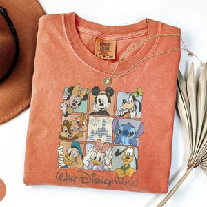 Może przedstawiać: Pomarańczowa koszulka Comfort Colors z kolorową grafiką postaci Disneya, w tym Myszki Miki i Myszki Minnie oraz Goofy'ego. Koszulka ma okrągły dekolt, a pod grafiką nadrukowano napis "Walt Disney World". Złoty naszyjnik i brązowy kapelusz r&oacute;wnież znajdują się na zdjęciu.