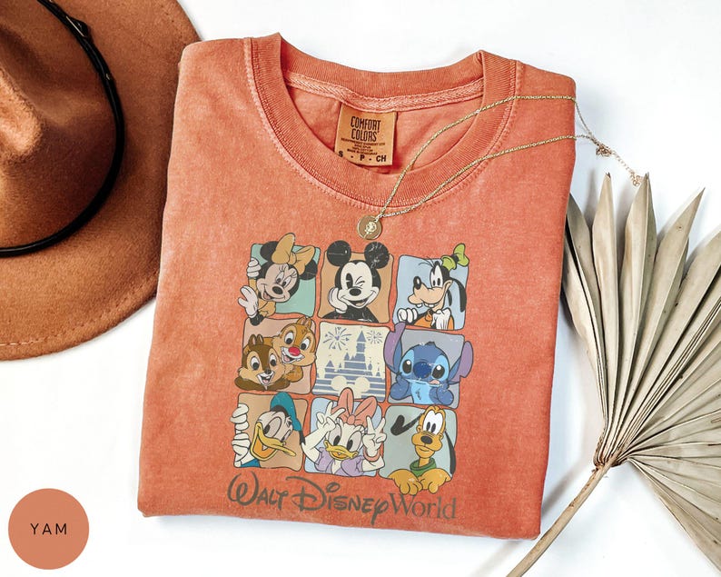 Może przedstawiać: Pomarańczowa koszulka Comfort Colors z kolorową grafiką postaci Disneya, w tym Myszki Miki i Myszki Minnie oraz Goofy'ego. Koszulka ma okrągły dekolt, a pod grafiką nadrukowano napis "Walt Disney World". Złoty naszyjnik i brązowy kapelusz r&oacute;wnież znajdują się na zdjęciu.
