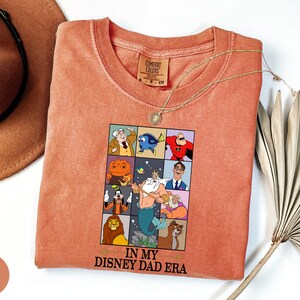 May include: Coral-colored t-shirt featuring a graphic of Disney characters in a grid, with the text "IN MY DISNEY DAD ERA." The shirt has a crew neck and short sleeves. A gold necklace is draped across the shirt. A brown hat is in the background.