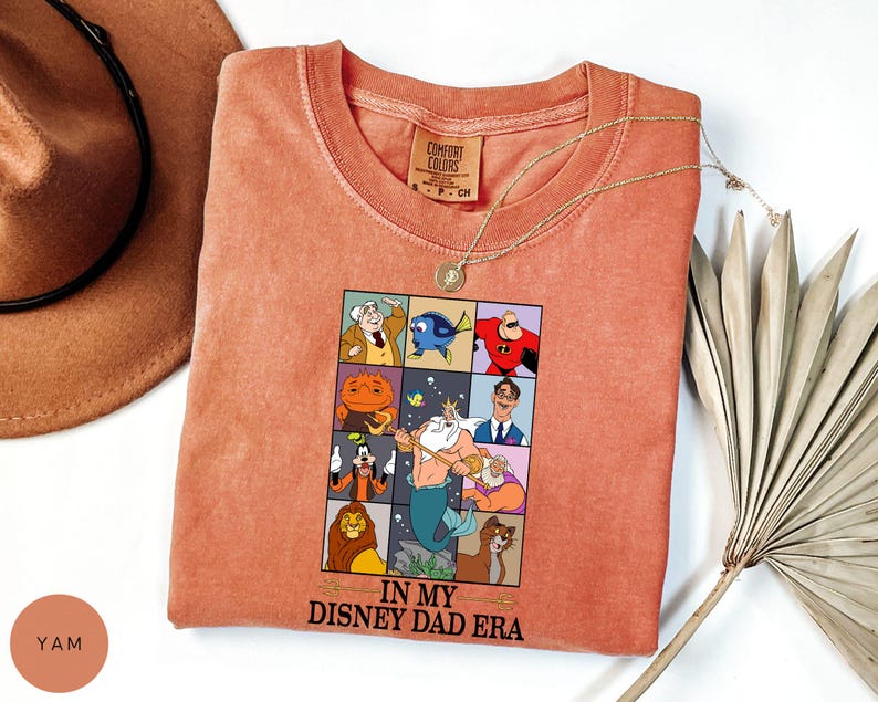 May include: Coral-colored t-shirt featuring a graphic of Disney characters in a grid, with the text "IN MY DISNEY DAD ERA." The shirt has a crew neck and short sleeves. A gold necklace is draped across the shirt. A brown hat is in the background.