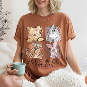 May include: A brown t-shirt featuring four cartoon characters: Winnie the Pooh, Eeyore, Tigger, and Piglet. The shirt has the text "POOH & CO. EST. 1926" printed below the characters. The shirt is worn by a person.