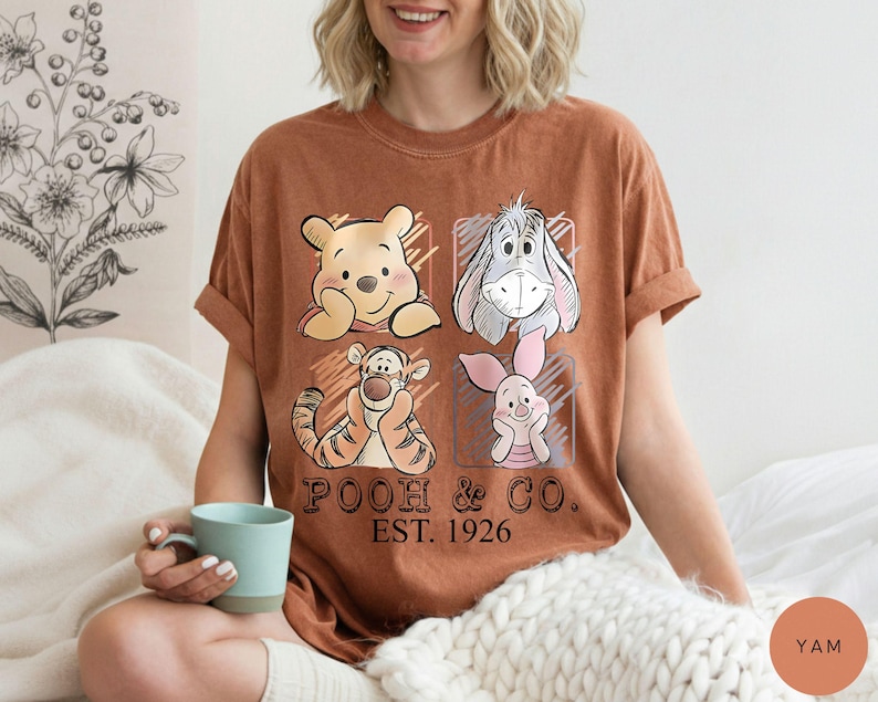 May include: A brown t-shirt featuring four cartoon characters: Winnie the Pooh, Eeyore, Tigger, and Piglet. The shirt has the text "POOH & CO. EST. 1926" printed below the characters. The shirt is worn by a person.