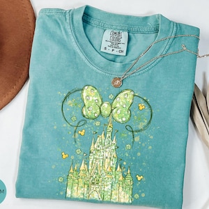 K&ouml;nnte beinhalten: T&uuml;rkisgr&uuml;nes T-Shirt mit einer Grafik eines Schlosses, einer Schleife und Mickey Mouse-Symbolen. Die Schleife ist hellgr&uuml;n mit wei&szlig;en Punkten. Eine goldene Halskette liegt &uuml;ber dem Shirt. Das Shirt ist mit "Comfort Colors" gekennzeichnet.