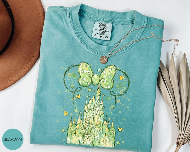 K&ouml;nnte beinhalten: T&uuml;rkisgr&uuml;nes T-Shirt mit einer Grafik eines Schlosses, einer Schleife und Mickey Mouse-Symbolen. Die Schleife ist hellgr&uuml;n mit wei&szlig;en Punkten. Eine goldene Halskette liegt &uuml;ber dem Shirt. Das Shirt ist mit "Comfort Colors" gekennzeichnet.