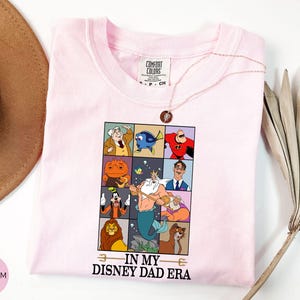 May include: A light pink t-shirt featuring a grid of Disney character illustrations. The shirt has the phrase "IN MY DISNEY DAD ERA" printed below the grid. A gold necklace with a pendant is draped across the shirt.