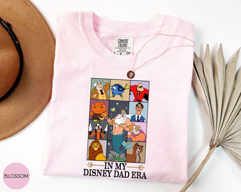 May include: A light pink t-shirt featuring a grid of Disney character illustrations. The shirt has the phrase "IN MY DISNEY DAD ERA" printed below the grid. A gold necklace with a pendant is draped across the shirt.