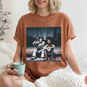 May include: A rust-colored short-sleeve t-shirt featuring a graphic print of two animated characters sitting on a curb at night. One character is a space ranger in a white and green suit, and the other is a cowboy wearing a hat and vest.