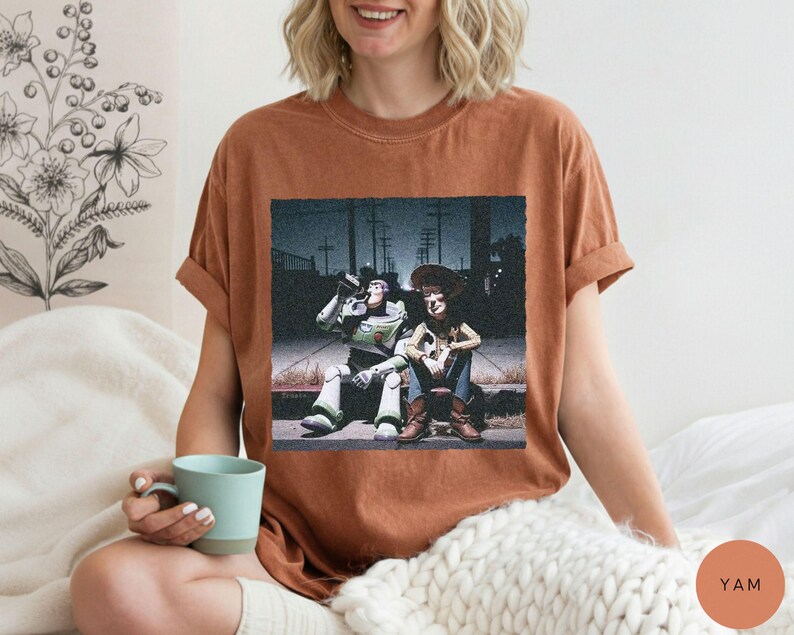 May include: A rust-colored short-sleeve t-shirt featuring a graphic print of two animated characters sitting on a curb at night. One character is a space ranger in a white and green suit, and the other is a cowboy wearing a hat and vest.