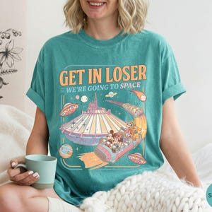 Pode incluir: Camiseta verde-&aacute;gua com um gr&aacute;fico retr&ocirc; apresentando um passeio de parque tem&aacute;tico com tema espacial e o texto "GET IN LOSER WE'RE GOING TO SPACE". O design inclui personagens de desenhos animados e planetas.