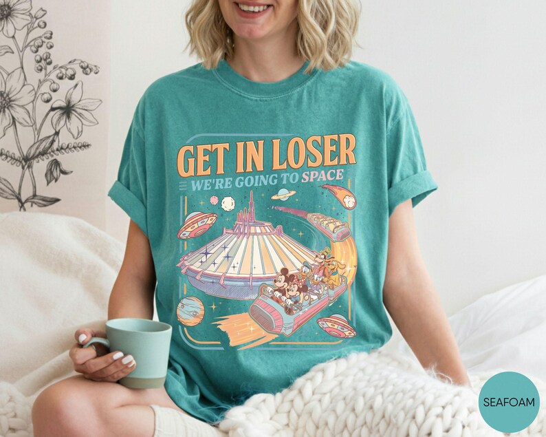 Pode incluir: Camiseta verde-&aacute;gua com um gr&aacute;fico retr&ocirc; apresentando um passeio de parque tem&aacute;tico com tema espacial e o texto "GET IN LOSER WE'RE GOING TO SPACE". O design inclui personagens de desenhos animados e planetas.
