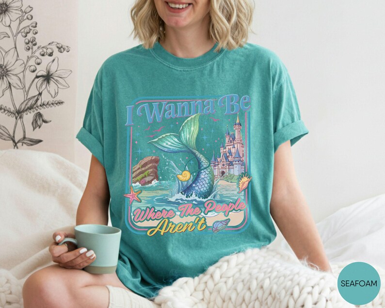 May include: Seafoam green t-shirt with a mermaid graphic and the text "I Wanna Be Where The People Aren't". The graphic includes a mermaid tail, castle, and ocean elements. The shirt is a comfortable, casual style.
