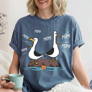 May include: A blue t-shirt with a cartoon design of two seagulls shouting "MOM!" and a small orange fish. The seagulls are white and black, standing on a rock. The shirt is a casual style, in a blue jean colour.