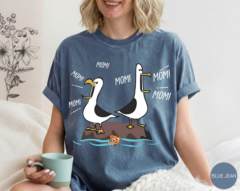 May include: A blue t-shirt with a cartoon design of two seagulls shouting "MOM!" and a small orange fish. The seagulls are white and black, standing on a rock. The shirt is a casual style, in a blue jean colour.