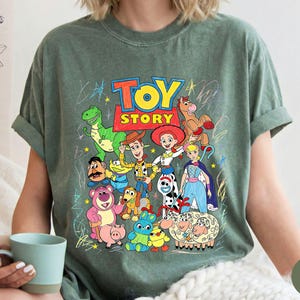 May include: A moss green short-sleeve t-shirt featuring a colourful graphic print of Toy Story characters including Woody, Jessie, Buzz Lightyear, Rex, and others. The design includes the Toy Story logo and scribbled star and line accents.